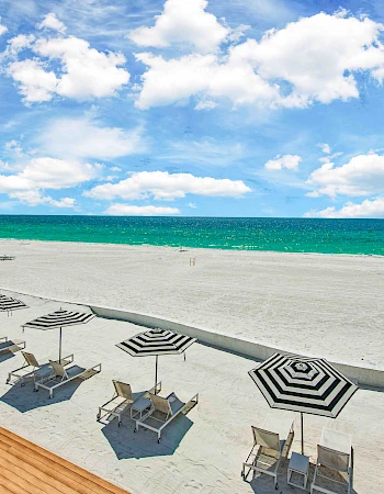 A beach with striped umbrellas and lounge chairs on sand, clear blue sky with clouds above, and a vibrant turquoise sea in the background.