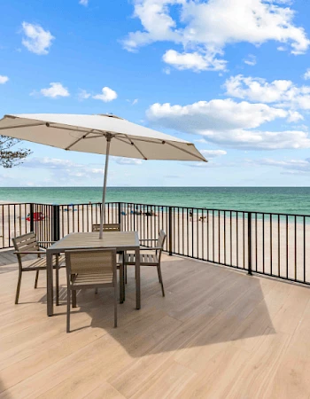 A beachside deck with a table, chairs, and umbrella overlooks the ocean. A perfect spot for relaxation under a blue sky with clouds.