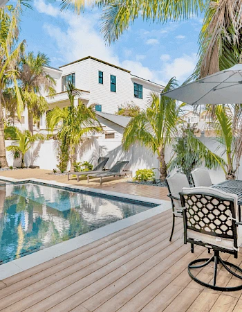 A luxurious outdoor space with a pool, surrounded by palm trees and a patio with dining furniture under an umbrella.