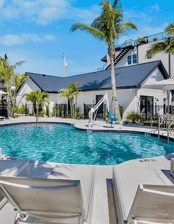 A luxurious outdoor pool area with sun loungers, parasols, palm trees, and modern buildings in the background, under a clear blue sky.