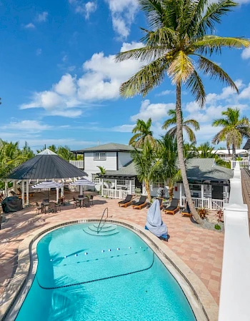 A serene outdoor scene with a swimming pool, palm trees, and a patio area featuring tables and chairs, under a clear blue sky.