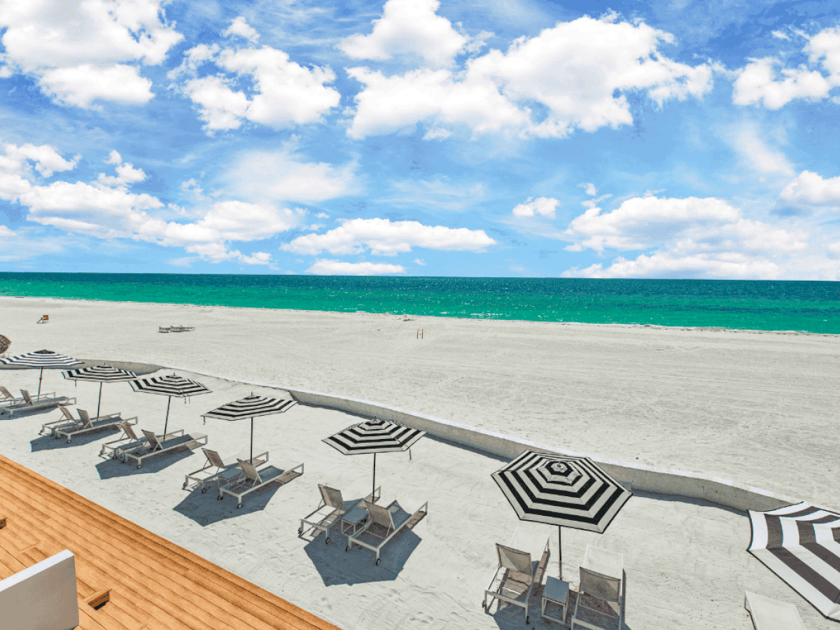 A beach scene with empty lounge chairs and umbrellas on the sand, overlooking a turquoise ocean under a blue sky with clouds.