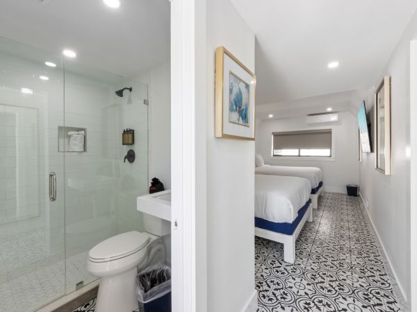 A modern hotel-style suite with a glass-enclosed bathroom on the left and a bright, tiled bedroom corridor with two beds on the right.