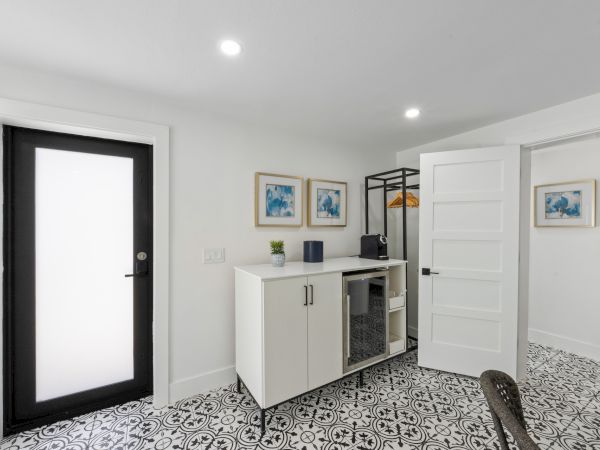 A bright, modern entry area with a black-framed glass door, white cabinetry, a small desk, framed art, and patterned tile flooring.