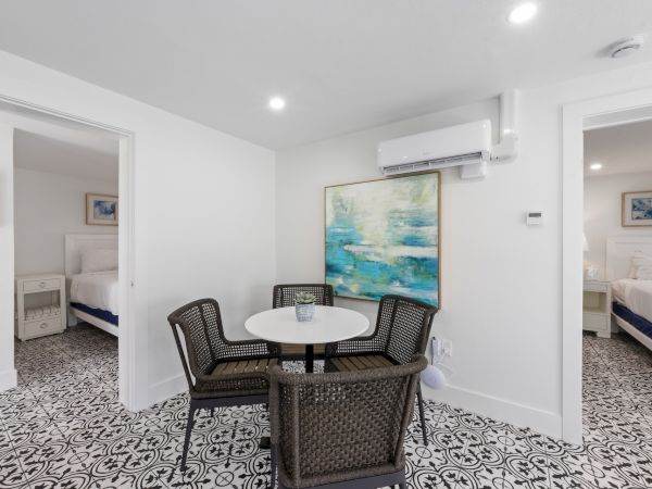 A bright dining nook with a round table and four wicker chairs, patterned tile floor, white walls, and two open doorways to bedrooms.