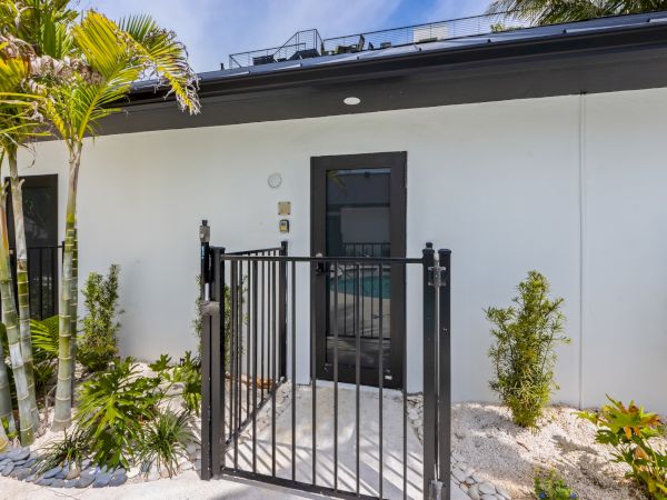 A modern single-story white house with a black metal gate and lush tropical plants lining the entry, small gravel yard, and a dark front door.