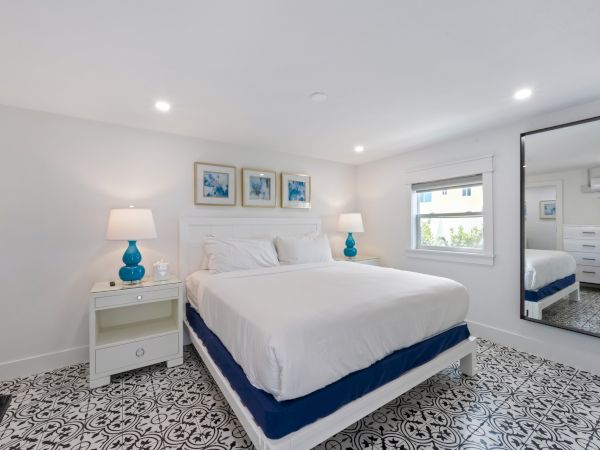 A bright, modern bedroom with a white-and-blue coastal theme, featuring a queen bed, matching nightstands, turquoise lamps, patterned tile floor, and a full-length mirror.