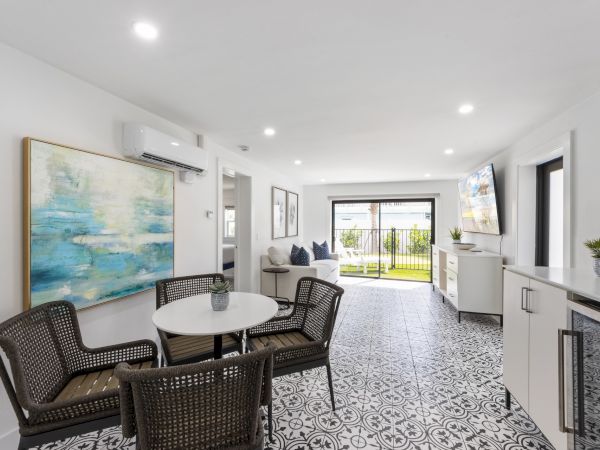 A bright, modern living area with a patterned tile floor, round dining table and wicker chairs, open kitchen, large glass doors to a backyard, and wall art.