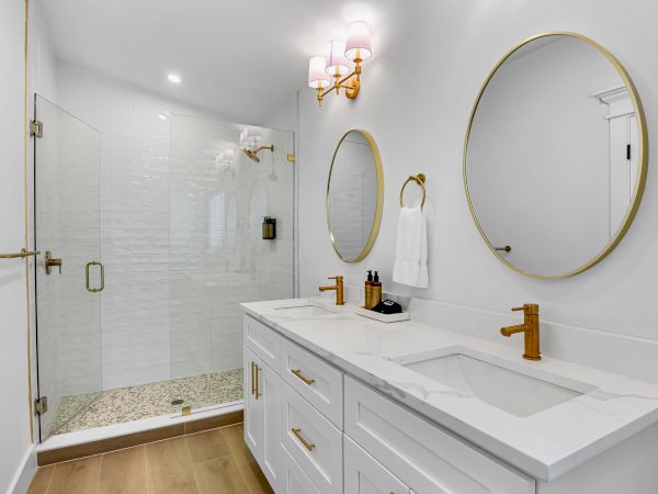 A stylish bathroom with a double vanity, white cabinetry, round mirrors, gold fixtures, and a glass enclosed shower with pebble floor.