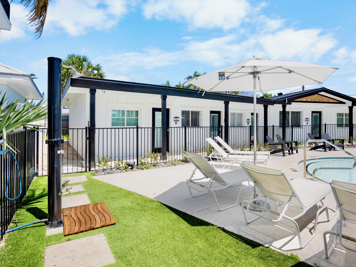 Pool area with lounge chairs, umbrellas, and modern apartments in the background. Bright and sunny day with blue skies.