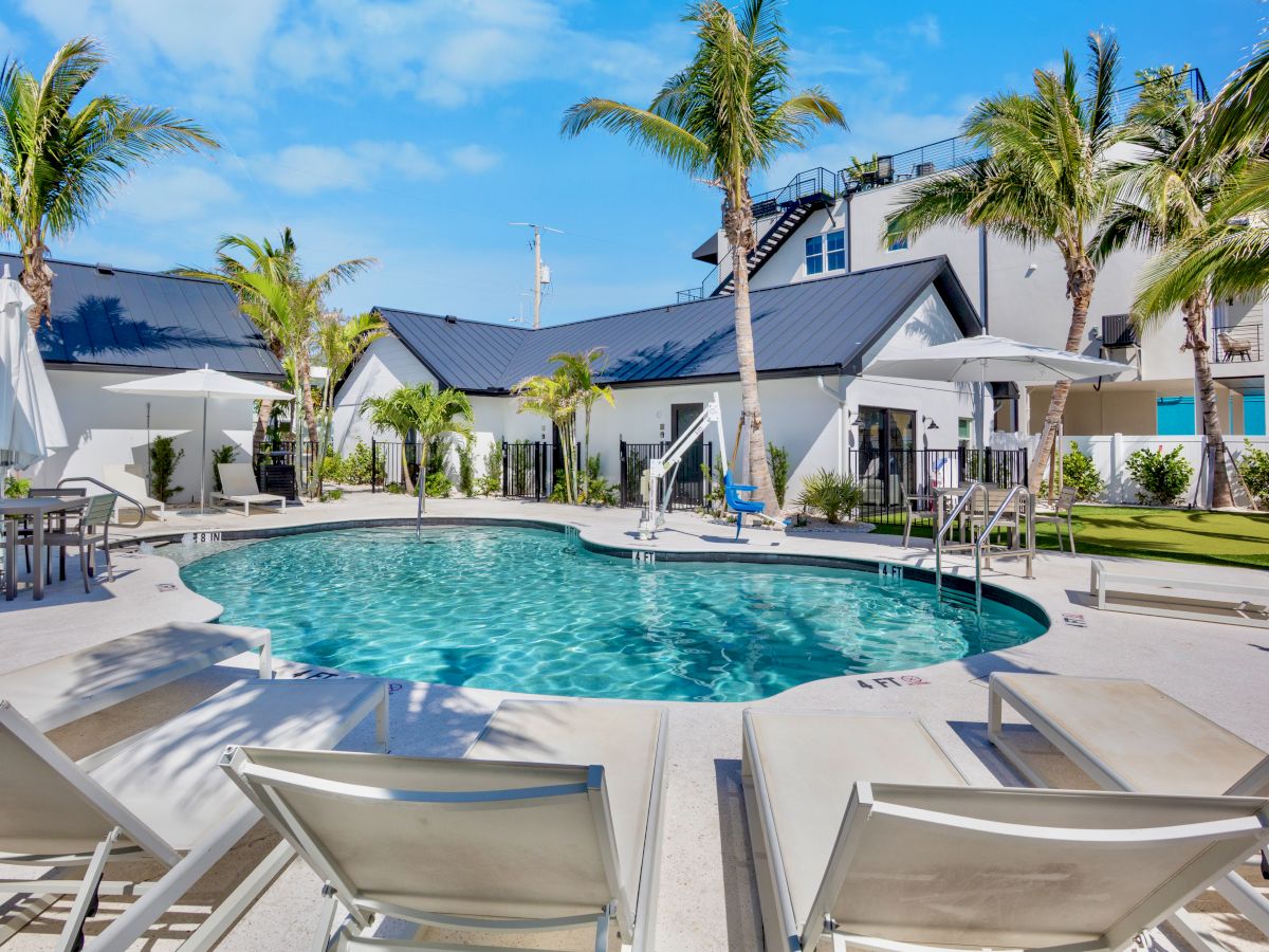 A luxurious outdoor pool area with sun loungers, umbrellas, and palm trees, surrounded by modern buildings under a clear blue sky.
