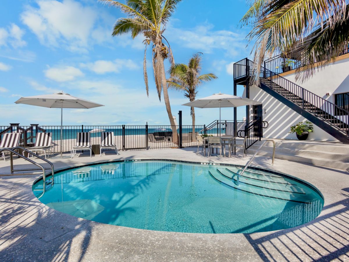 A sunny poolside scene with a circular pool, palm trees, lounge chairs, umbrellas, and a railing overlooking the sea, next to a white building.