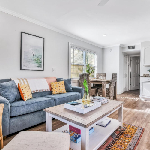 Bright living room with a blue sofa, coffee table, dining set by windows, and a white kitchen with stove and cabinets.