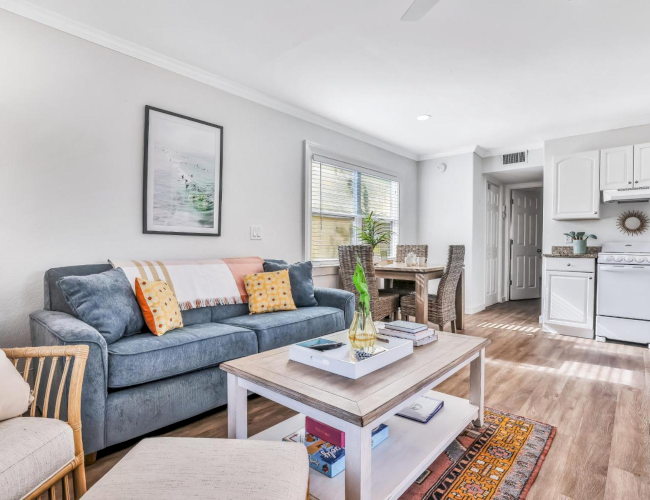 Bright living room with a blue sofa, coffee table, dining set by windows, and a white kitchen with stove and cabinets.