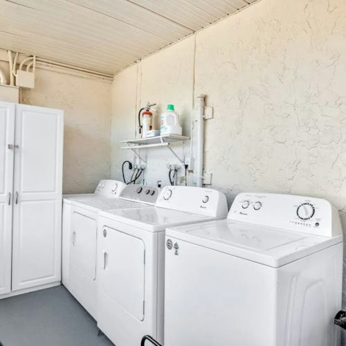 A clean laundry room shows two white washer-dryer machines with plumbing, shelves, and supplies against the wall.