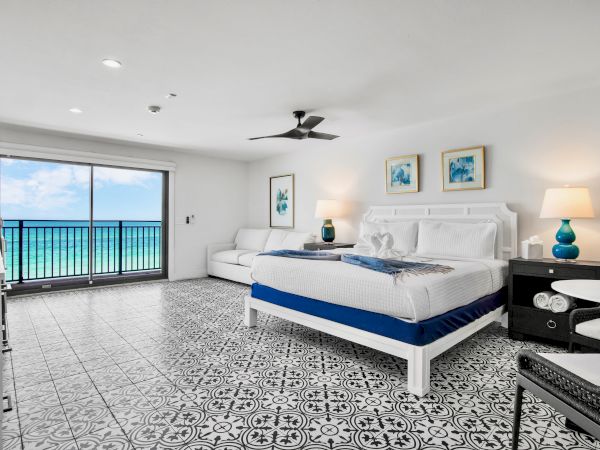 A bright modern bedroom with a white bed, blue accents, ceiling fan, framed art, and a large ocean-view balcony door.