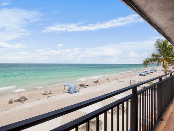 View from a balcony overlooking a sandy beach with palm trees, sun umbrellas, and calm turquoise ocean under a cloudy blue sky.
