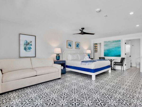 Modern bedroom with a white sofa and king bed, blue lamps, ceiling fan, framed artwork, and patterned tile floors.