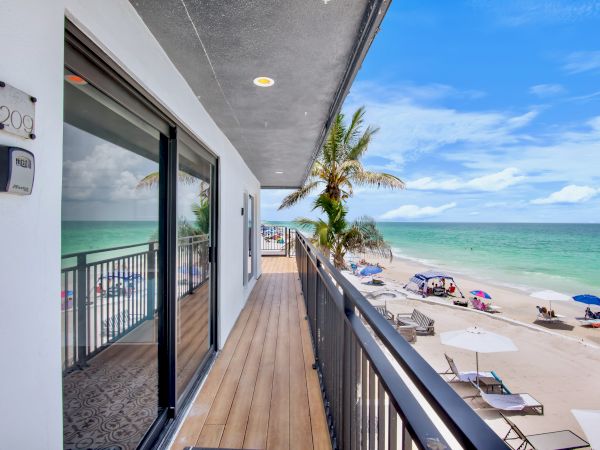 View from a hotel balcony with sliding doors, palm trees, and sun loungers along a turquoise beach under a blue sky.