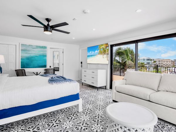 Bright modern bedroom with a ceiling fan, bed and sofa, and large doors opening to a sunny palm-lined beach view.