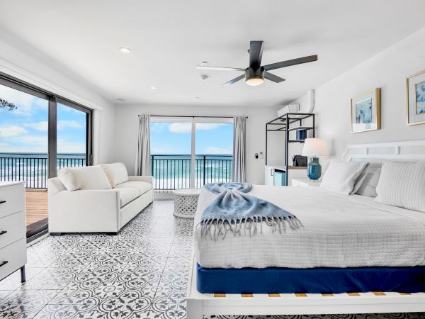 Bright modern bedroom with an ocean-view balcony, white sofa and bed, a black ceiling fan, and framed blue artwork.