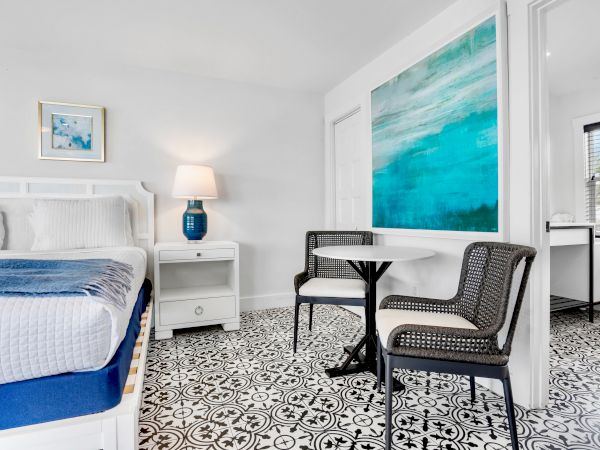 A bright bedroom sitting area with a bed, lamp, small table, patterned chairs, and large blue abstract artwork on the wall.