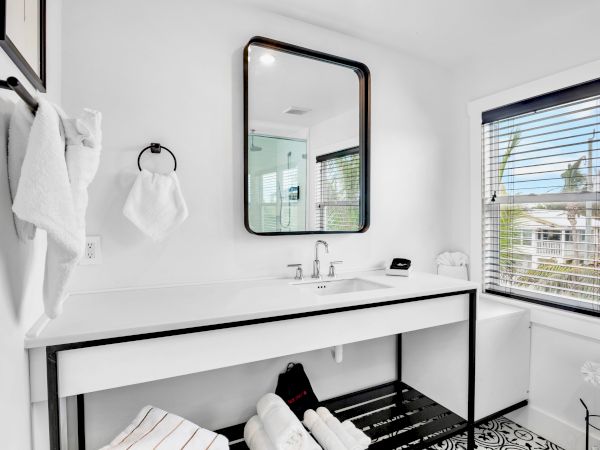 Bright modern bathroom with a white vanity, framed mirror, towel hooks, and a window with blinds.