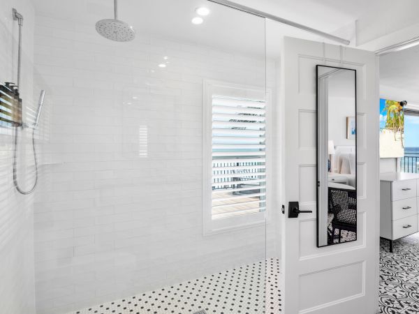 A modern bathroom shower with a rainfall head, white tile walls, and a vanity area opening to a sunny ocean view.