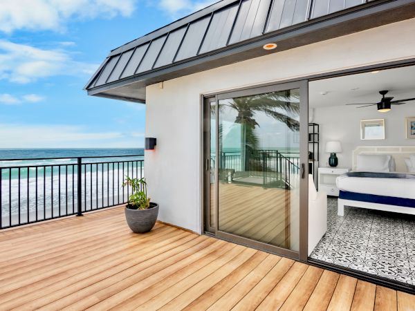 Ocean-view balcony with wooden deck, railing, and an open modern bedroom through sliding glass doors.