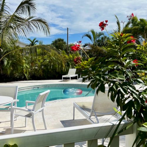A sunny tropical pool with lounge chairs, red flowers, and palm trees under a blue sky with clouds.