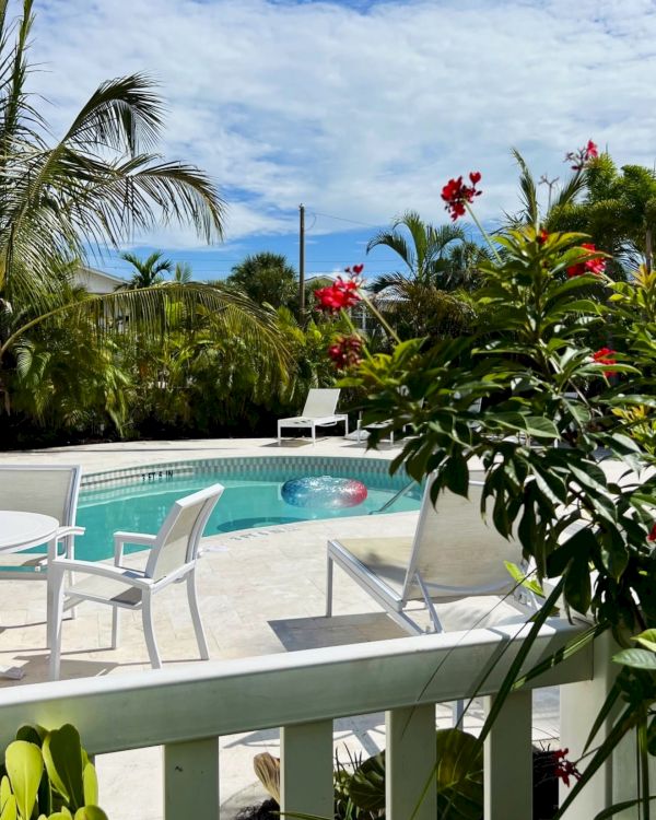 A sunny tropical pool with lounge chairs, red flowers, and palm trees under a blue sky with clouds.