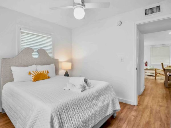 A bright bedroom with a queen bed, table lamp, ceiling fan, and an open door to a small hallway.