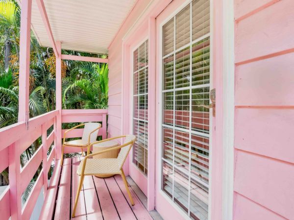 A pastel pink balcony with wicker chairs and glass louvered shutters overlooking lush palm trees.