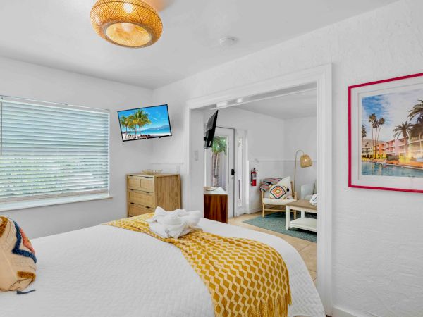 Bright bedroom with a bed, white blinds, a wall TV, a ceiling light, and a framed tropical pool photo.
