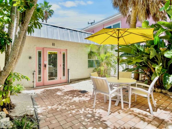 Tropical courtyard with pink doors, a yellow patio umbrella, white chairs and table, and lush palms and trees.