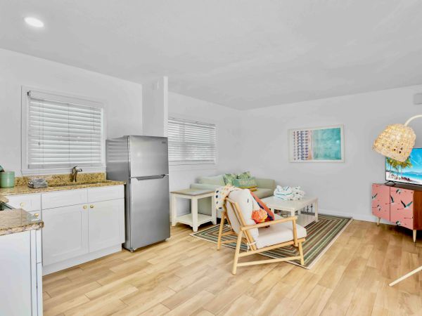 A bright open-plan room with a kitchen counter, refrigerator, couch, TV stand, and kids chair near wall art.