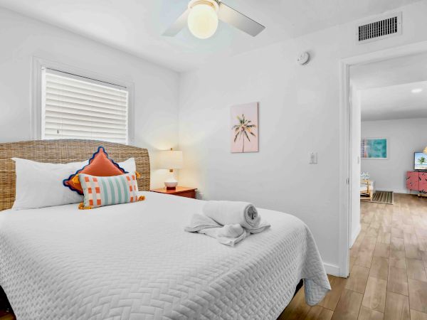 A bright bedroom with a wicker bed, ceiling fan, window blinds, beach-themed art, and an open doorway to a TV room.