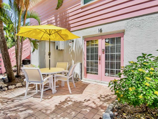 Sunny patio with white chairs and table under a yellow umbrella, beside a pink door and barred window, with palm trees.