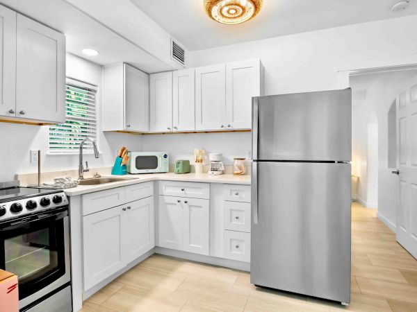Bright white kitchen with cabinets, sink, oven, refrigerator, toaster, and microwave by a window with blinds.