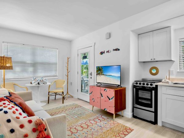 Bright living room-kitchen with a sofa, TV showing a beach, an island with a decorative chest, a gas stove, cabinets, and blinds.