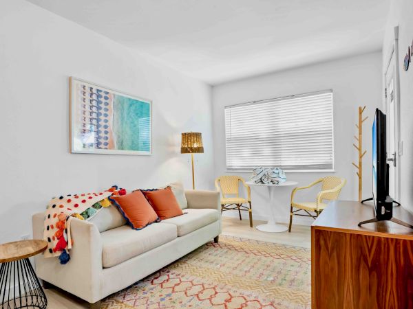 Bright living room with a sofa, colorful pillows, framed wall art, window blinds, two chairs, and a standing TV.