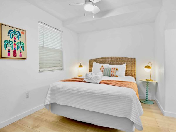 A bright bedroom with a wicker headboard bed, white bedding and pillows, framed art, window blinds, ceiling fan, and two bedside lamps.