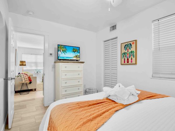 A bright bedroom with a TV on a dresser, framed artwork, white shutters, and an orange-accented bed with towels.