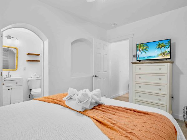 A bright hotel bedroom with a bed, dresser, and wall TV showing a tropical beach, with an attached bathroom visible.