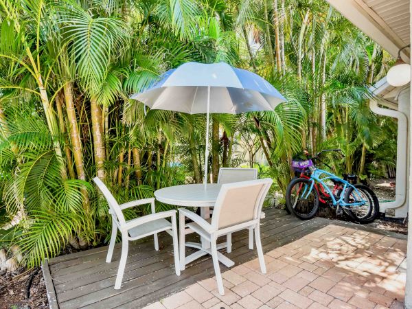 A sunny patio with a blue umbrella over white table and chairs, lush palm greenery, and bicycles near the wall.