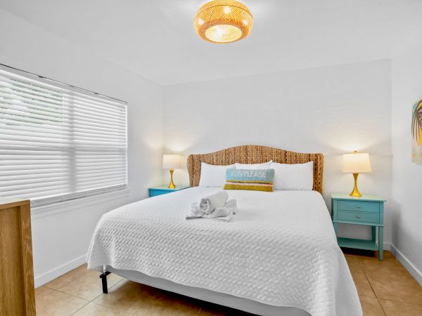 A bright bedroom with a neatly made bed, woven headboard, bedside lamps, teal cabinets, blinds, and a ceiling light.