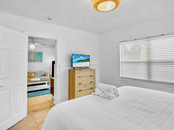 A bright bedroom with a neatly made bed, a TV on a wooden dresser, window blinds, and a warm ceiling light.