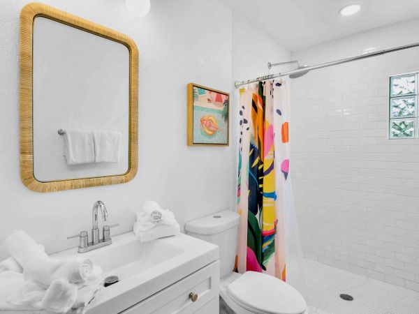 Bright bathroom with a wicker-framed mirror, white tub and toilet, colorful shower curtain, towels, and a sink.