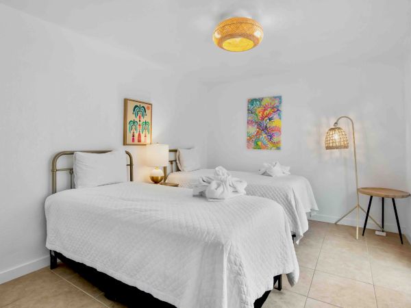 A bright bedroom with two white beds, a warm ceiling lamp, colorful wall art, a side table, and a woven floor lamp.