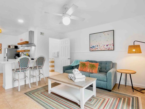 Bright open-plan living room/kitchen with blue sofa, coffee table, ceiling fan, framed art, and two white bar stools.